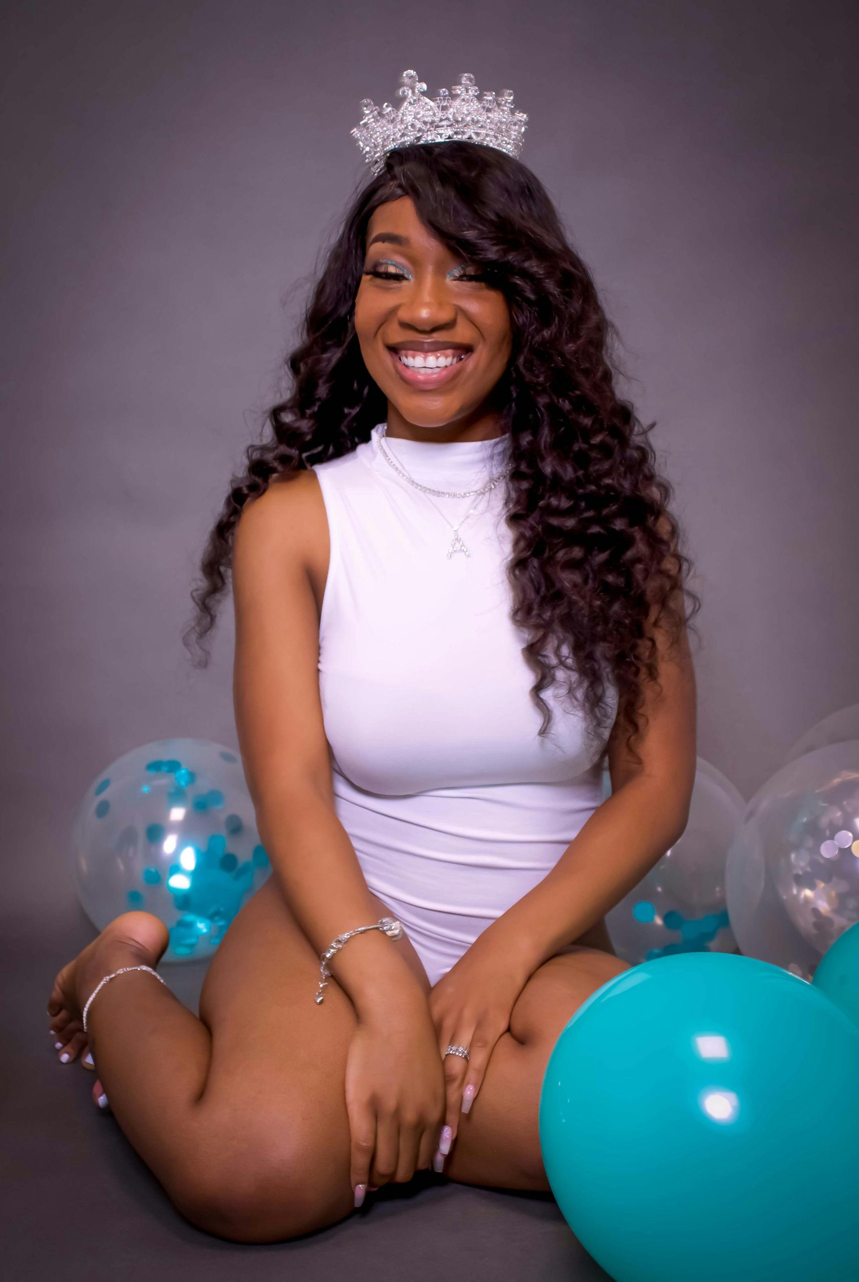 Smiling young woman in a white dress with a crown, surrounded by balloons, exuding joy.