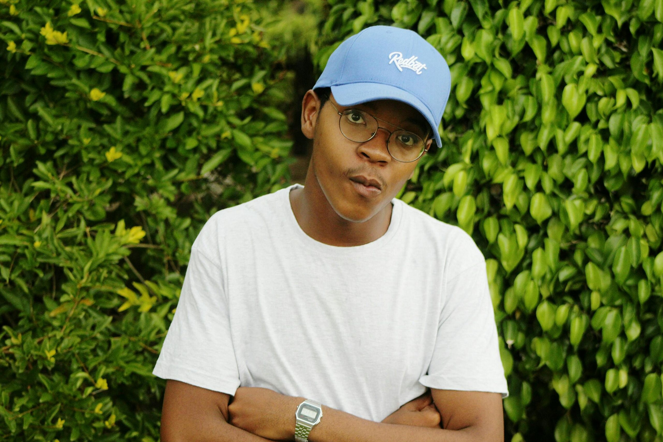 Young man in a white shirt and blue cap posing outdoors with greenery.